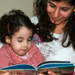 Jaël (Trisomie 18) mit 7 Jahren, auf dem Arm ihrer Mama, schaut sich ein Buch an, das ihre Mama hält.