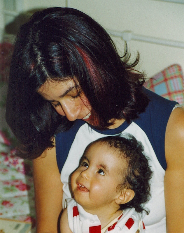 Die zweijährige Jaël (mit Trisomie 18) sitzt auf dem Schoß ihrer Mutter, die beiden lächeln sich an, während sich ihre Blicke treffen.